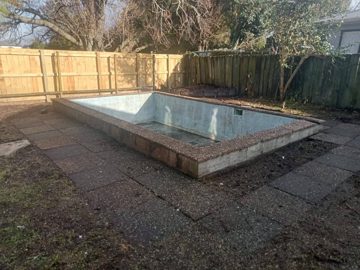 Empty swimming pool surrounded by paving in a fenced backyard.