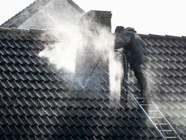 A worker washing a tiled roof with a pressure washer and ladder in place.