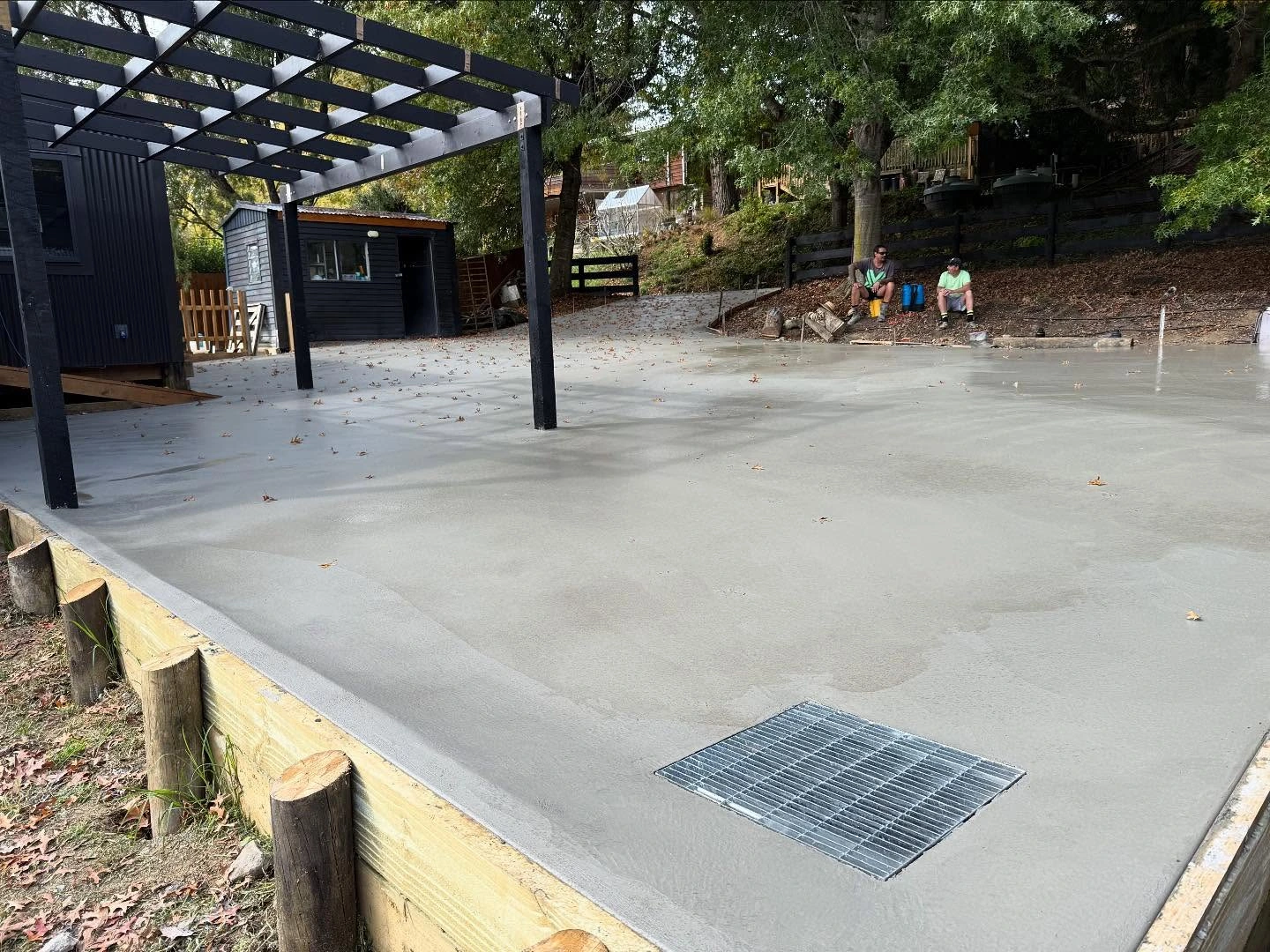 Smooth concrete patio beneath a black pergola, with timber edging and a drain
