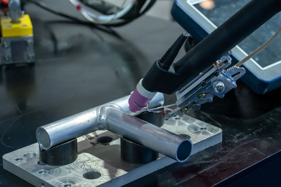A welding torch works along a joint between metal tubes held in a jig